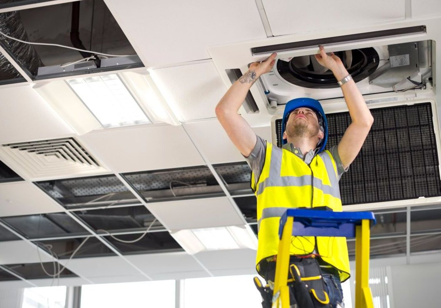 Image showing an electrician on a ladder fitting air conditioning unit into a ceiling. In a bright office an engineer in a hard hat stands on a ladder looking up as he fits an air conditioning unit into the ceiling.