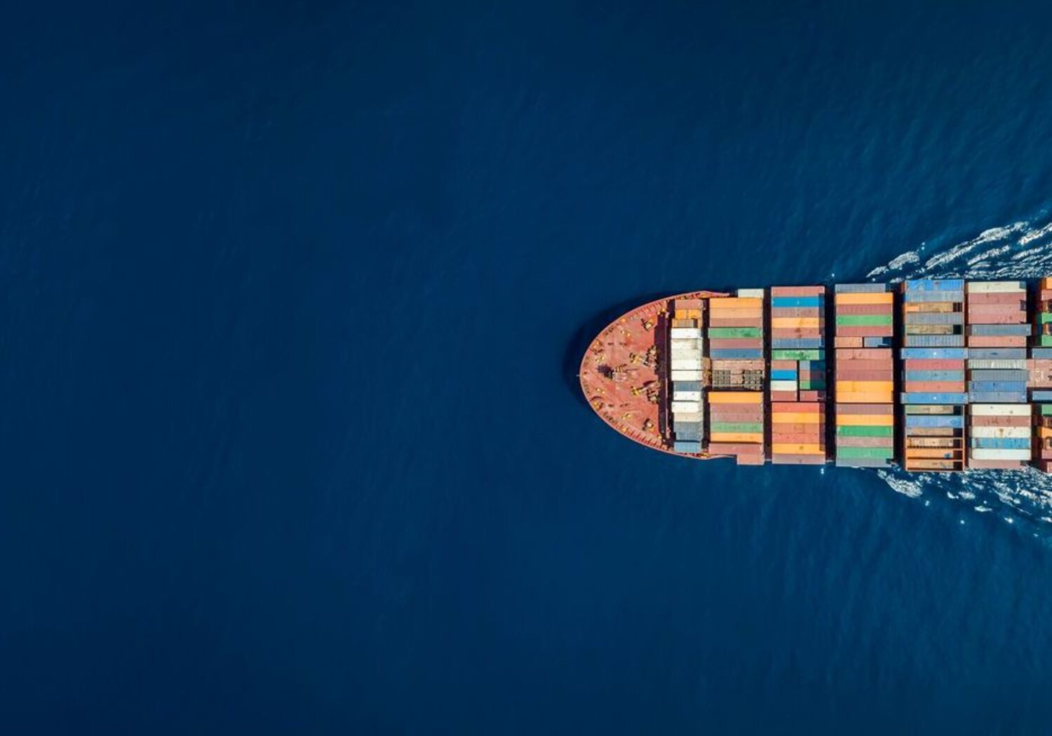 Image showing an aerial view of the top of a large cargo container ship.A picture from above of the surface of a dark blue sea.  A large cargo ship stacked with containers is emerging from the right hand side of the view.