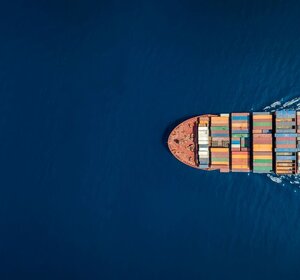 Image showing an aerial view of the top of a large cargo container ship.A picture from above of the surface of a dark blue sea.  A large cargo ship stacked with containers is emerging from the right hand side of the view.