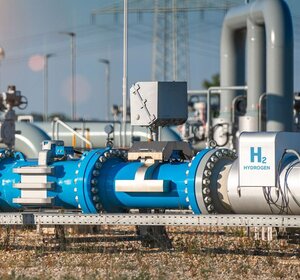 Image showing a pipeline from a hydrogen production plant. IN the foreground of a bright day is a silver and blue pipe from a hydrogen production plant with the words “H2” and “hydrogen” in large blue letters.