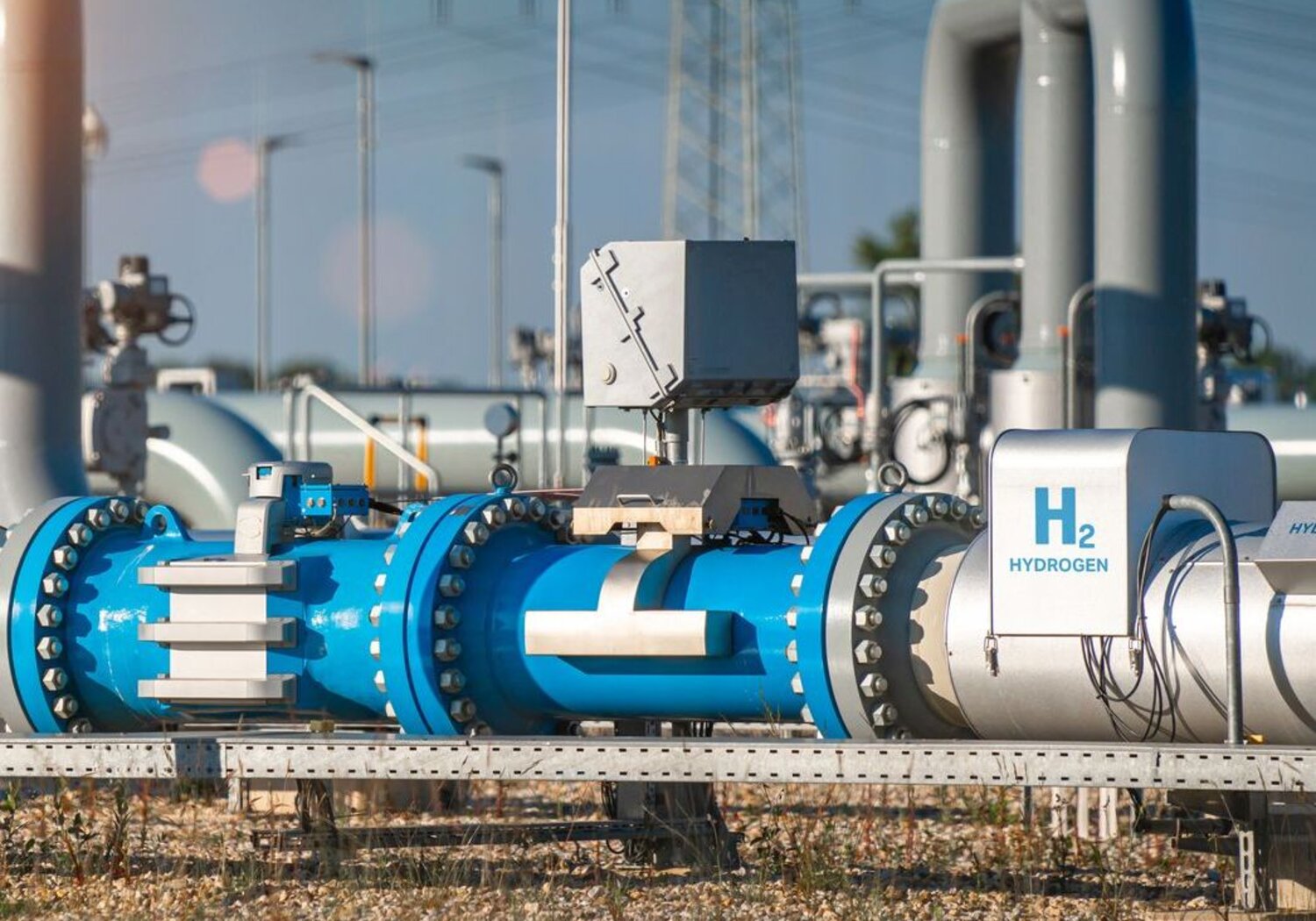 Image showing a pipeline from a hydrogen production plant. IN the foreground of a bright day is a silver and blue pipe from a hydrogen production plant with the words “H2” and “hydrogen” in large blue letters.