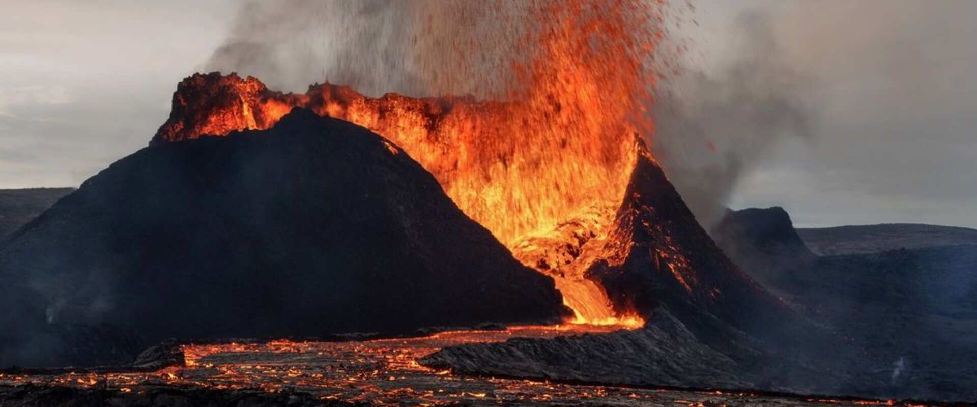 In the foreground is an erupting volcano in Iceland with a lava flow descending its flank