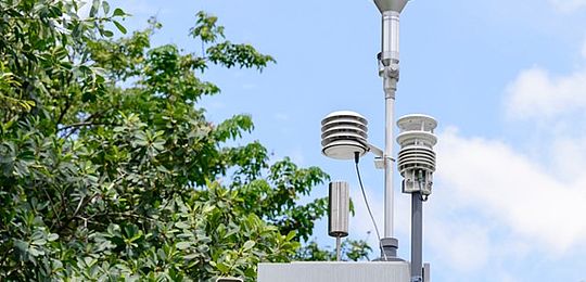 In the centre of the frame, an air monitoring station is attached to a pole. It consists of a grey metal box with a handle pointing towards the viewer and three covered pylons, carrying sampling equipment, protruding from the top, as well as a small silver cylinder. In the background there is the canopy of a full, green tree, and a blue sky filled with soft clouds