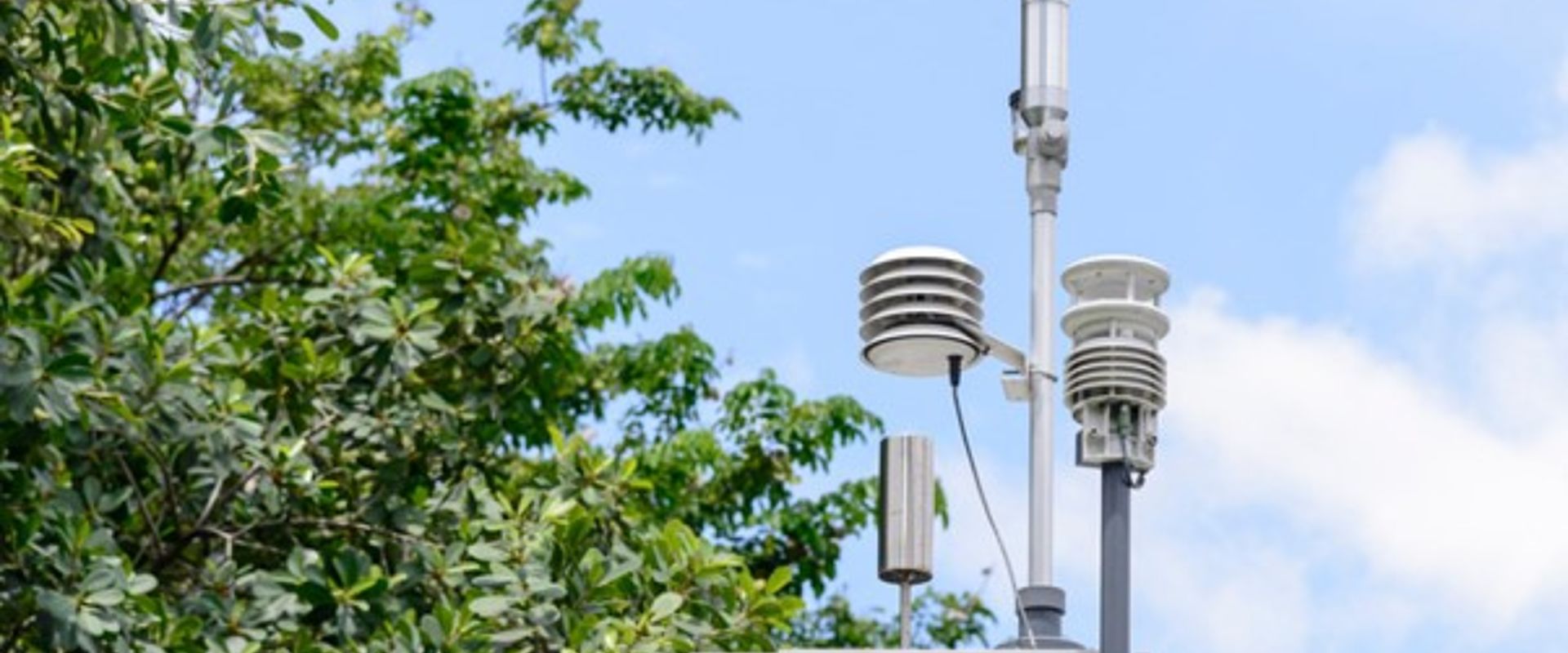 In the centre of the frame, an air monitoring station is attached to a pole. It consists of a grey metal box with a handle pointing towards the viewer and three covered pylons, carrying sampling equipment, protruding from the top, as well as a small silver cylinder. In the background there is the canopy of a full, green tree, and a blue sky filled with soft clouds