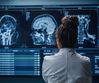 In the foreground is the back of a female neuroscientist in a white lab coat and in the background a wall of MRI scans of a patient’s brain that she is studying