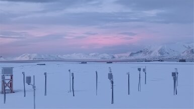 An image showing dawn over the Arctic research station in Norway. In the foreground measuring instruments stand out from the snow and in the background are a range of snow-covered mountains