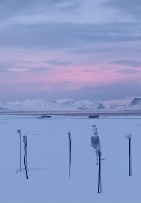 An image showing dawn over the Arctic research station in Norway. In the foreground measuring instruments stand out from the snow and in the background are a range of snow-covered mountains