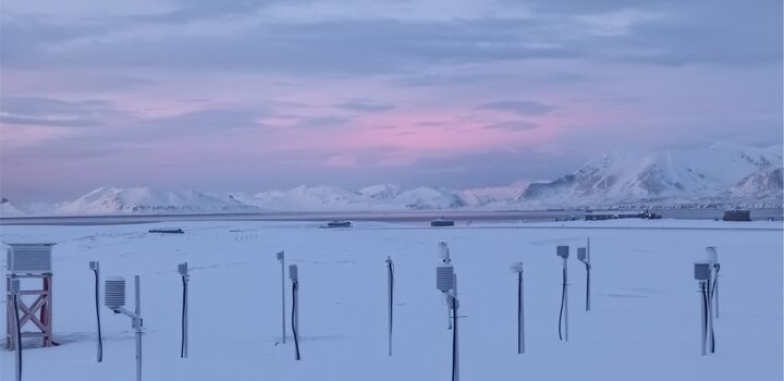 An image showing dawn over the Arctic research station in Norway. In the foreground measuring instruments stand out from the snow and in the background are a range of snow-covered mountains